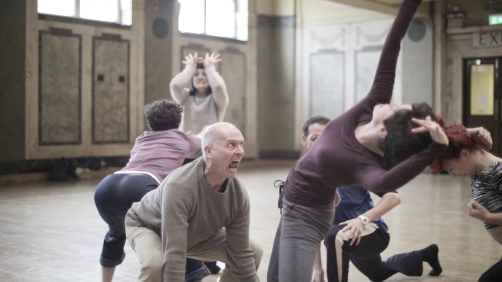A man crouches and shouts as dancers twist and stretch around him in an ornate rehearsal hall, from Nobody’s Guests