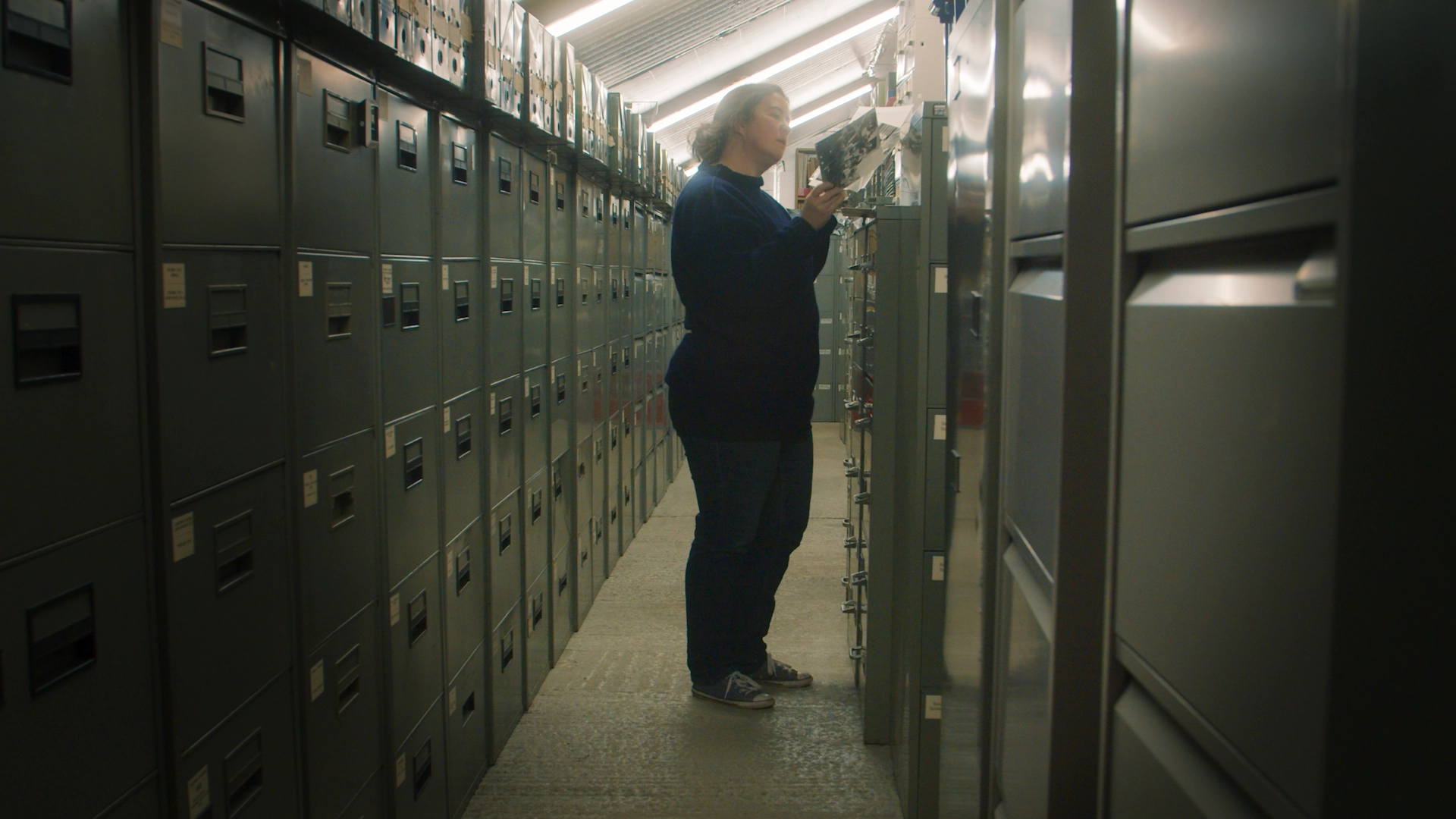 A woman studying a photograph in an archive aisle lined with metal filing cabinets, from The Moment of Truth